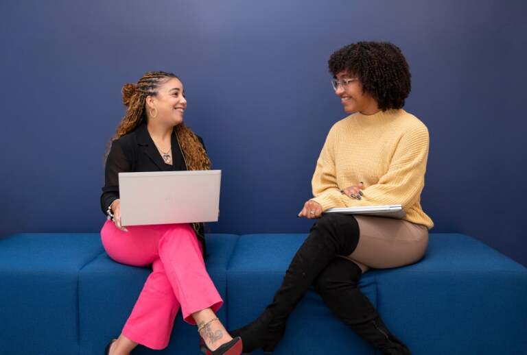 Two women sitting on a covered bench looking at each other and holding laptops.